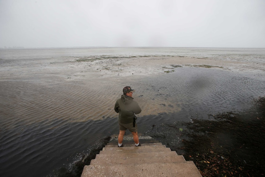 A man stands at the bottom of a set of concrete stairs and looks out at a bay where water has receded
