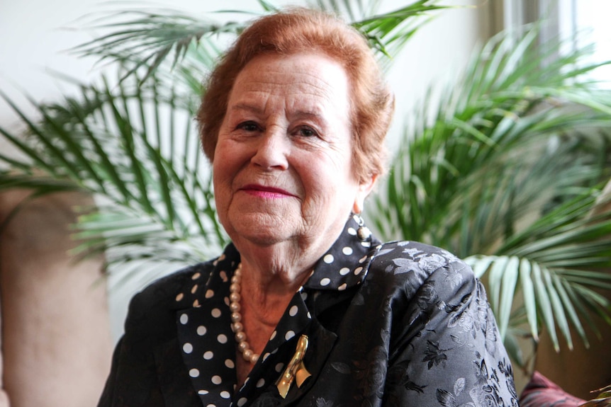 Elderly Jewish woman Yelena Gorodetsky, sits in formal attire. Indoor palm in background.