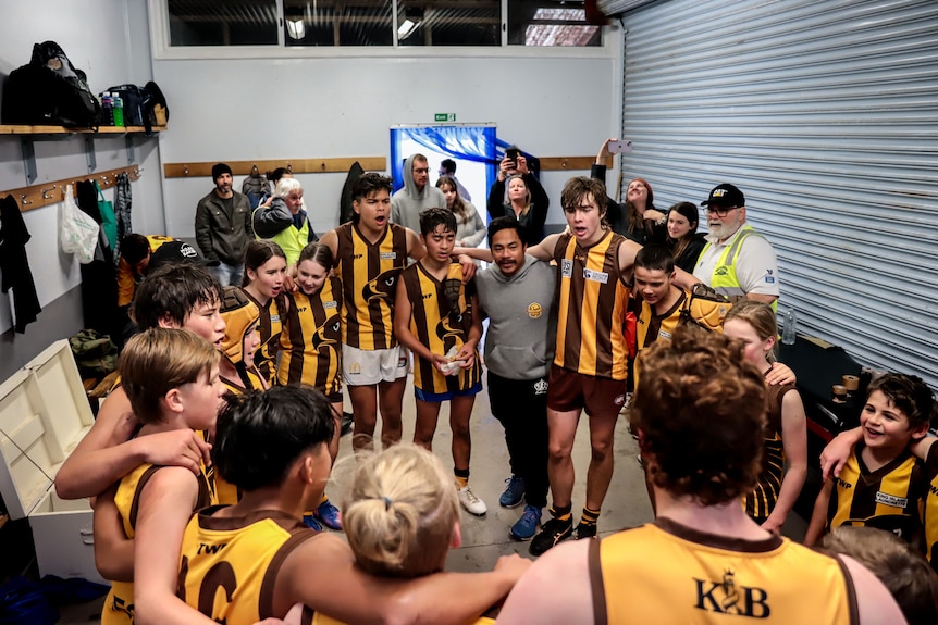 A circle of different aged teenagers wearing brown and gold football uniforms sing inside a changeroom 