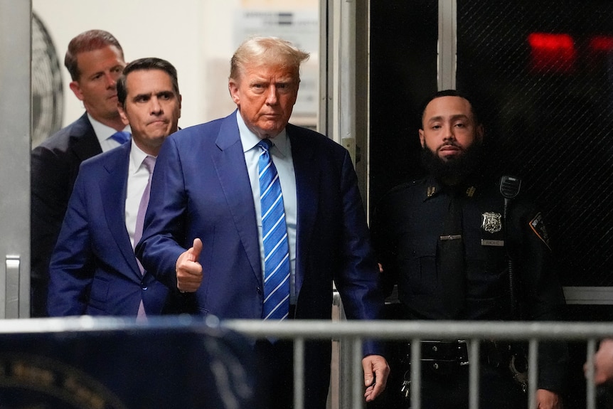 Donald Trump walking in a blue suit giving a thumbs up with a security guard on his left and two other men on his right