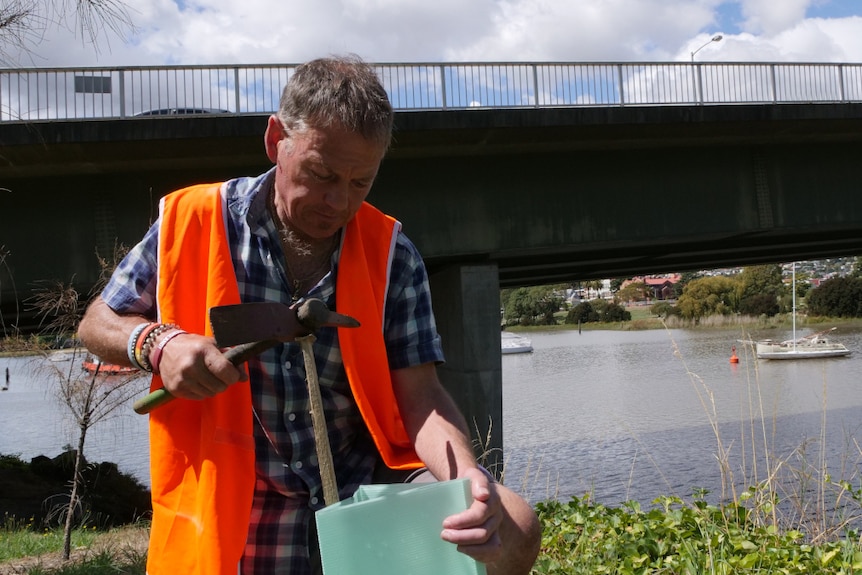 A man, wearing a high viz vest, plants a tree beside a river