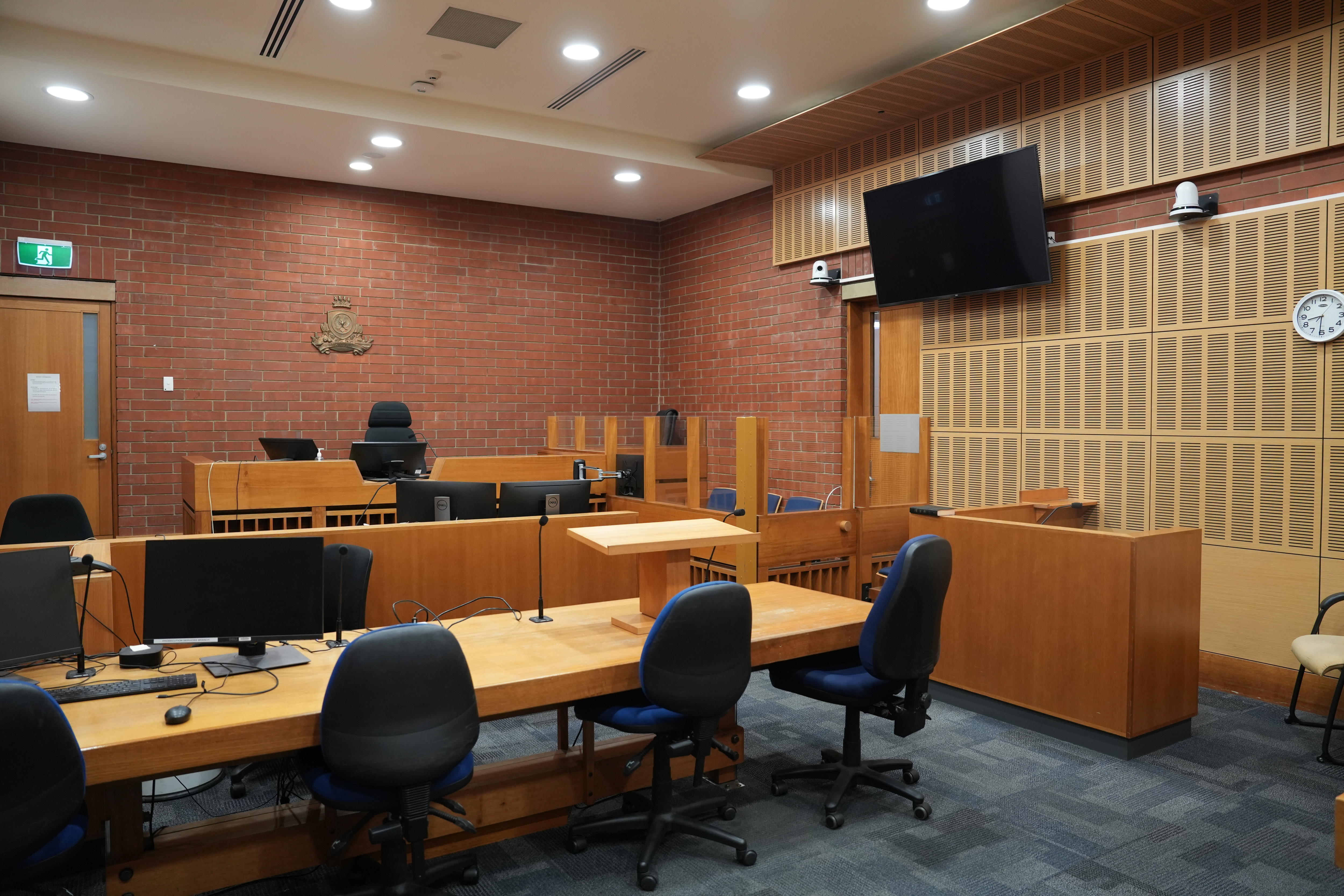 Escritorios y sillas vacíos frente al juez. Una pantalla de televisión cuelga de la pared sobre la zona del acusado, que está vallada por una pared de medio cristal.