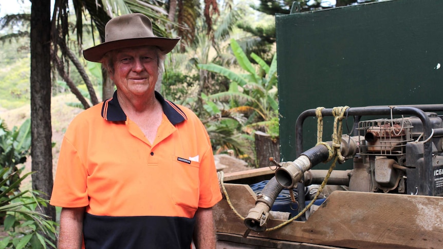 Senior man wearing high-vis shirt and felt bush hat stands next to truck carrying water pump