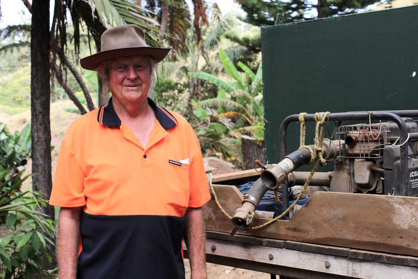 Senior man wearing high-vis shirt and felt bush hat stands next to truck carrying water pump