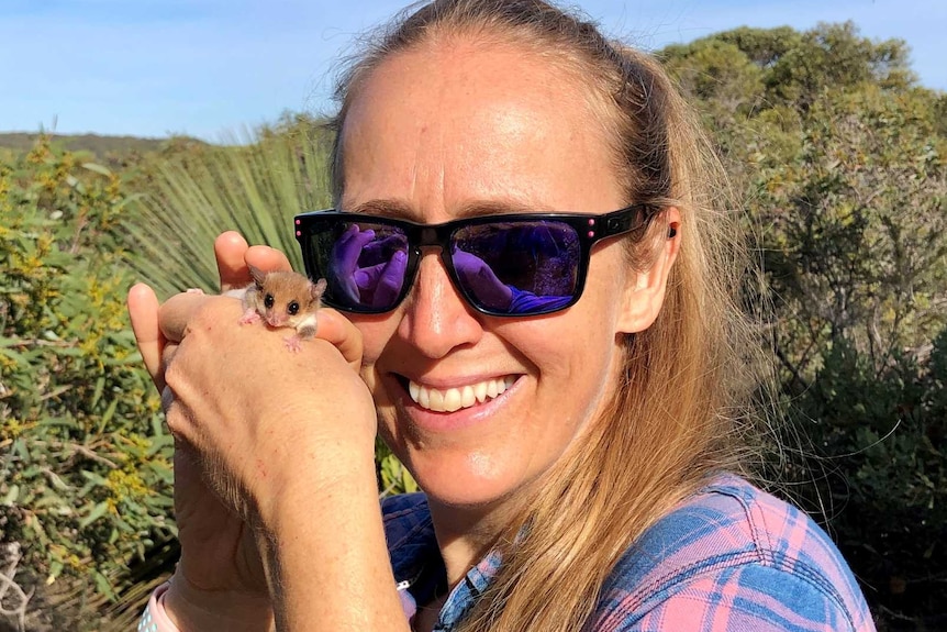 Woman wearing sunglasses in bush holding tiny mouse-like marsupial up to her face and looking at camera