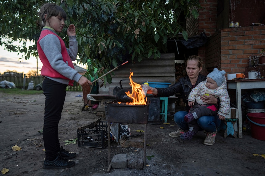 A woman with a baby holds a baby bottle over a fire outside, while another child holds a burning stick over the flame.