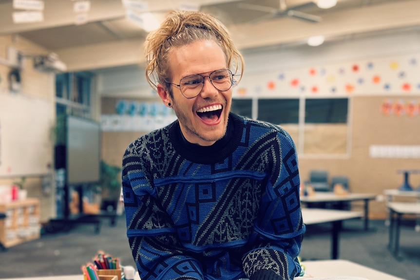 A man with a bun, glasses and knitted jumper sits on a table in a classroom.