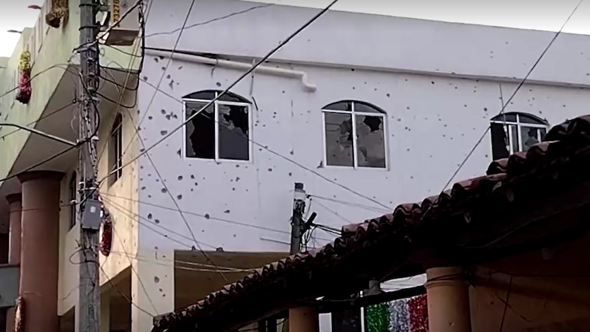 Bullet holes leave glass shattered in the three visable windows of a white building in the streets of Mexico.
