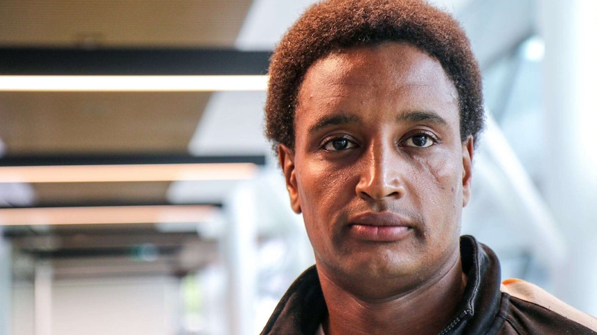 An African-Australian man looks at the camera, with light shining on his face.