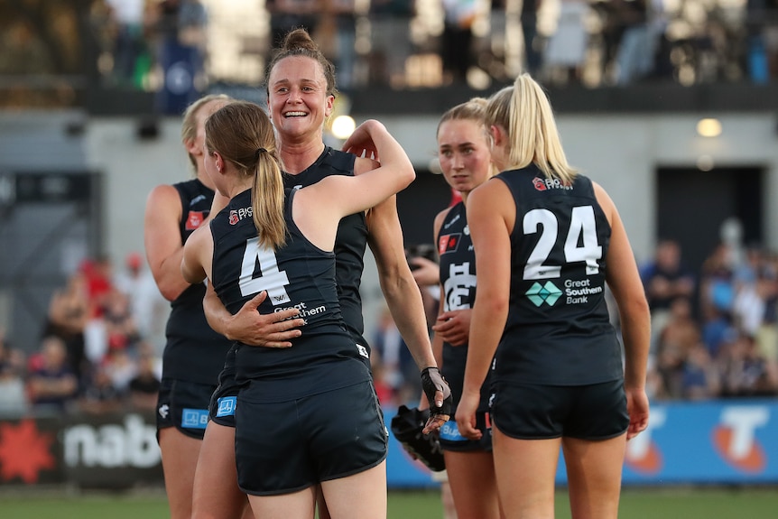 A group of Carlton AFLW players clad in navy blue grin and hug on the ground after a win.