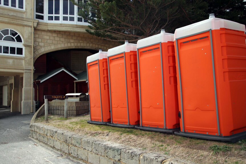 Four orange portable toilets on grass next to a building at Cottesloe Beach.