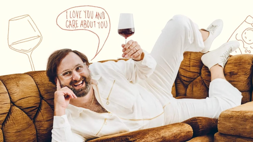 Eric Wareheim smiling, wearing a white jumpsuit, laying on a couch with a red wine in hand.