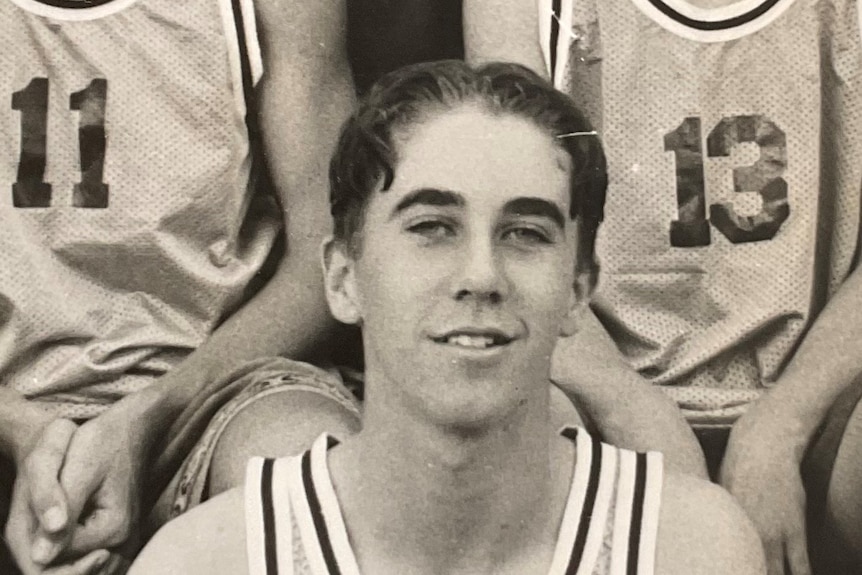 A black and white photo shows a young man smiling to the camera in a basketball jersey. 