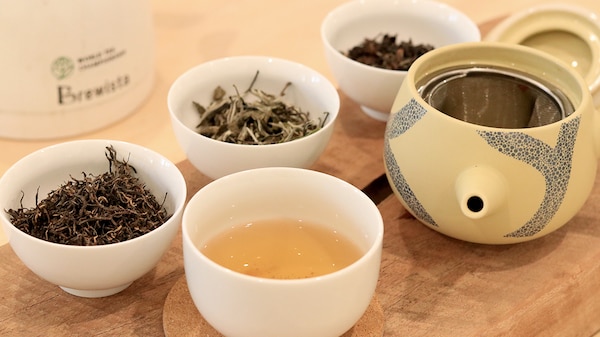 Tea leaves in small cups next to a cup of tea and a tea pot on a table.