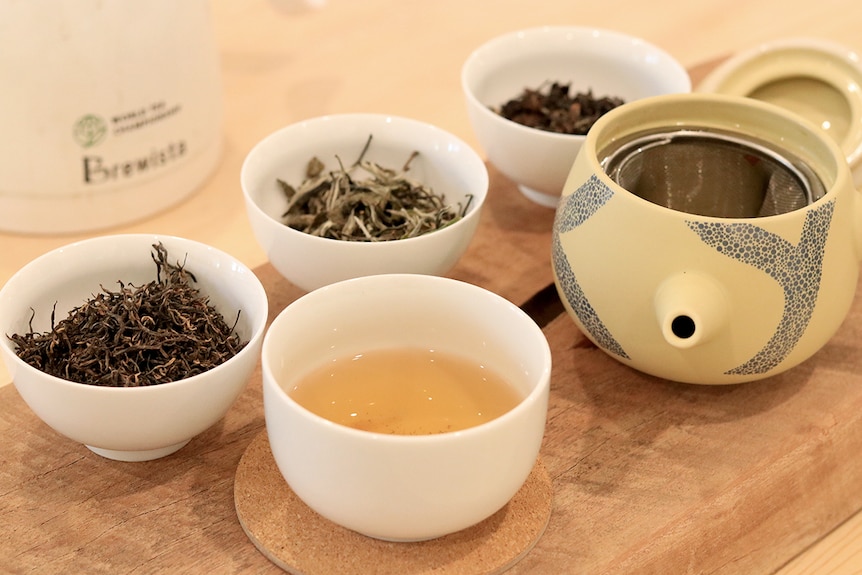 Tea leaves in small cups next to a cup of tea and a tea pot on a table.