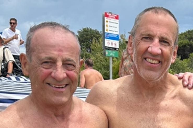 Two older men stand on the beach smiling.