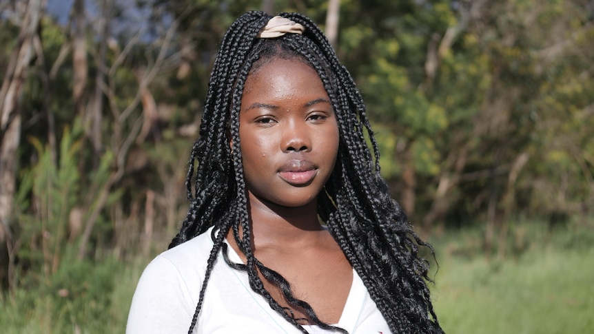 A teenage girl looks to the camera with a faint smile and smize in her eyes. She wears black box braids and a white top.