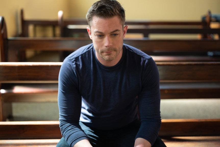 Tim Pocock sits on a church pew looking down. 