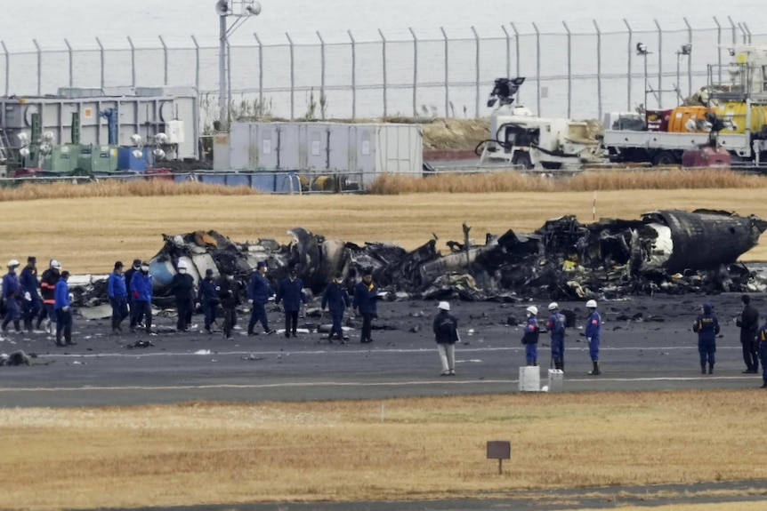 People in blue jumpsuits stand in front of a burnt-down aircraft on a runway 