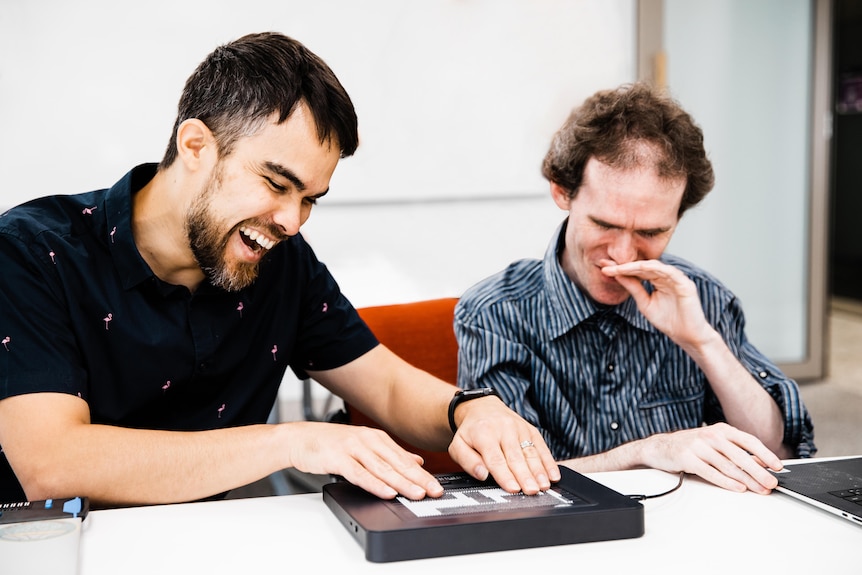 Jamie and Mick at a table, both have one hand on braille protoype, both looking down smiling. Mick has his hand over his mouth