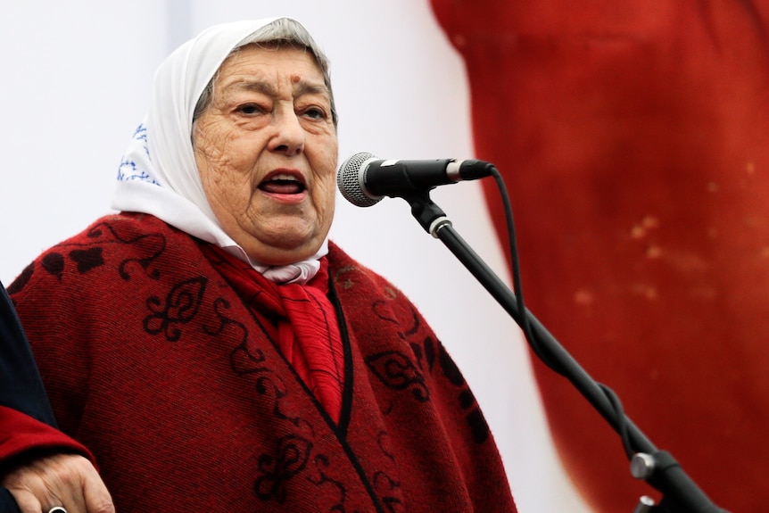 Hebe de Bonafini in a red cape and white headscarf speaks in front of a microphone. 