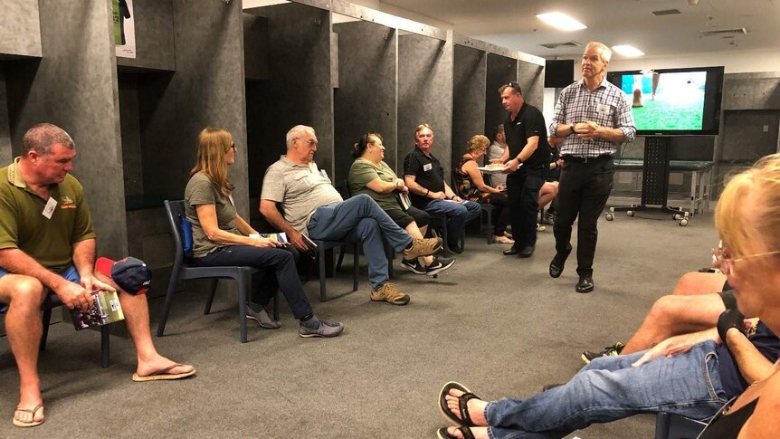 Matt Elliot stands in the middle of a changing room, talking to a group of men and women who are seated