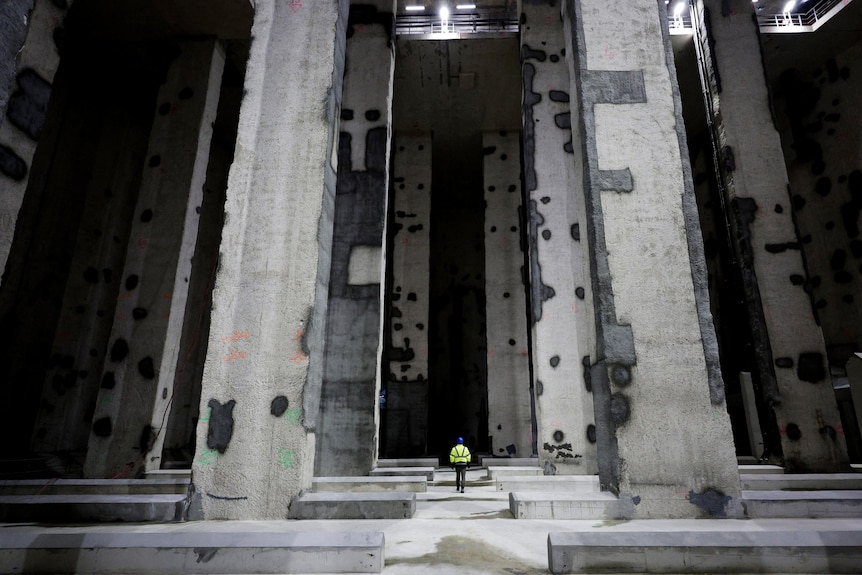 A man in a yellow hi-vis jacket is dwarfed by several large concrete pillars in a big empty space.