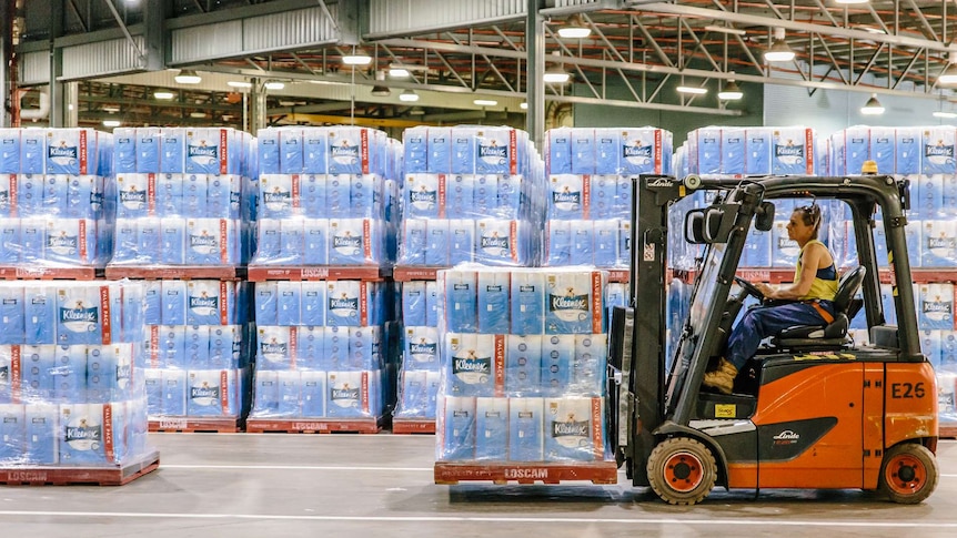 A woman drives a forklift inside a warehouse filled with toilet paper