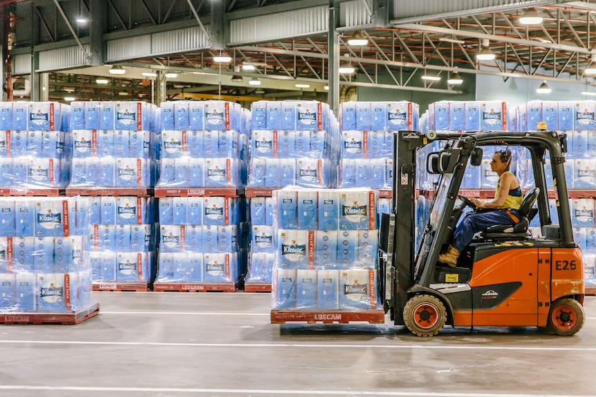 A woman drives a forklift inside a warehouse filled with toilet paper