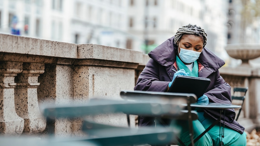 A woman wearing medical scrubs, a surgical face mask, gloves and a large puffer sits at a table outside writing on her notepad