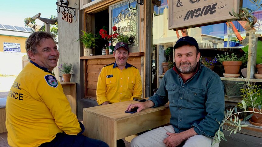 Three men sit at a cafe table. Two are in yellow RFS shirts and the other is in a blue long sleeve shirt.