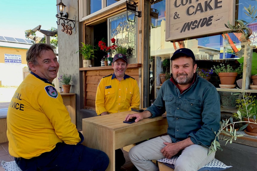 Three men sit at a cafe table. Two are in yellow RFS shirts and the other is in a blue long sleeve shirt.