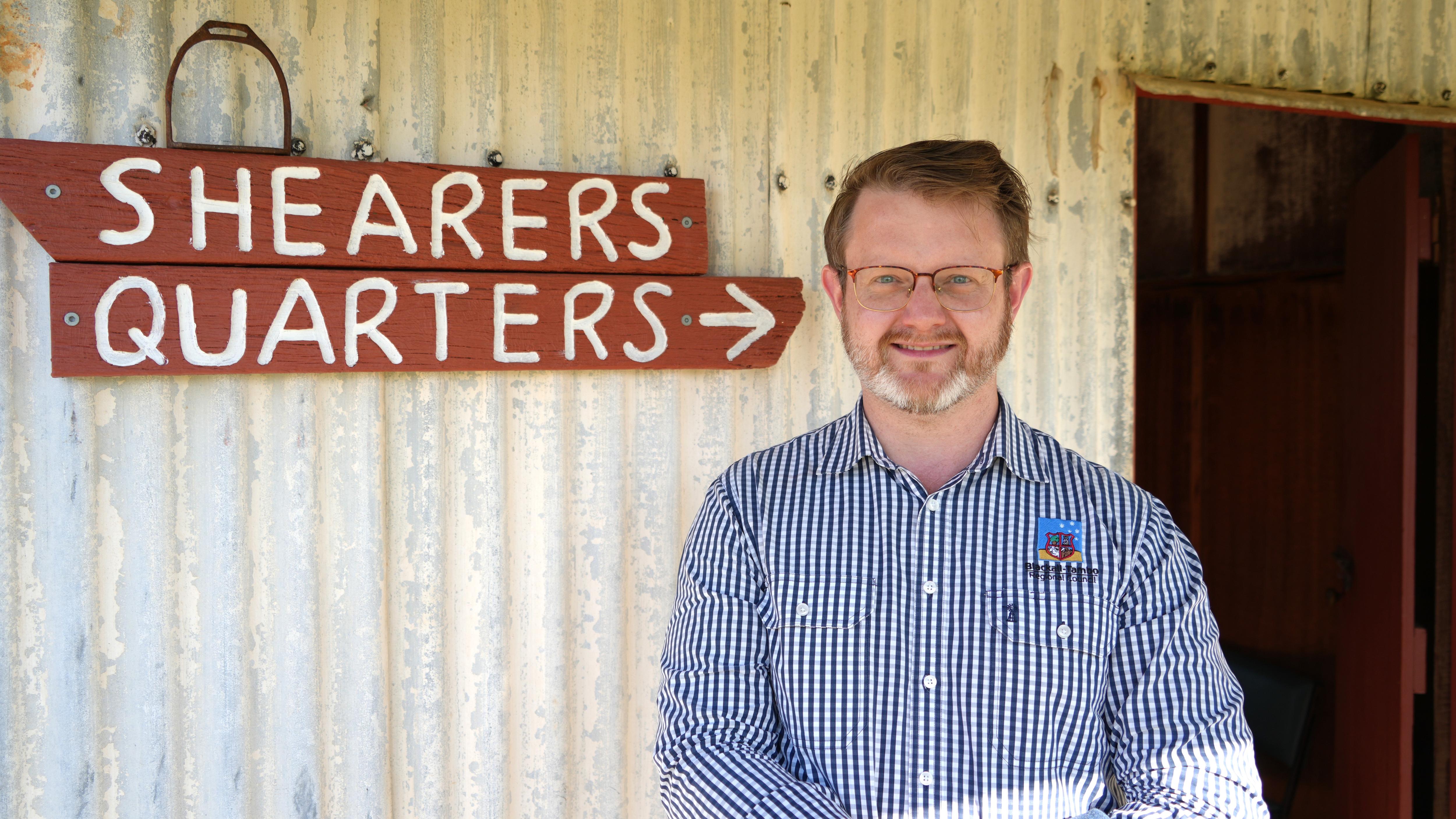A man with glasses poses in front of a corrugated iron wall on which a "Shearers quarters" sign hangs.