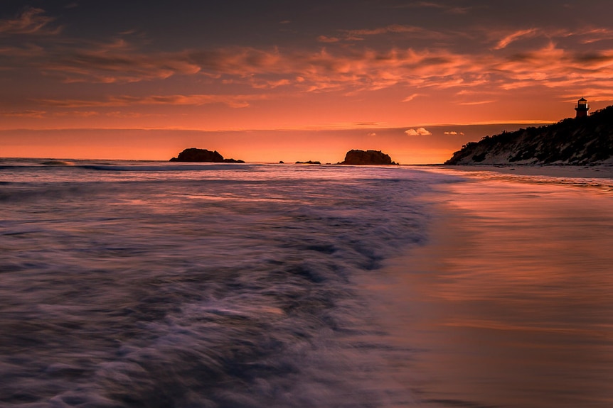A deep orange, yellow and pink sunset over calm crashing waves on a shore.
