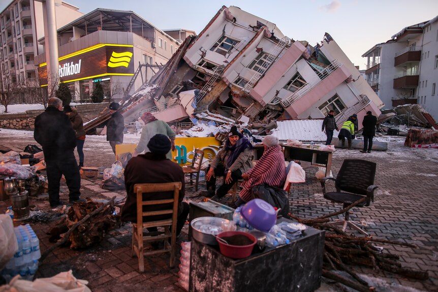 People stand by collapsed buildings, sitting at a table drinking tea under blankets.