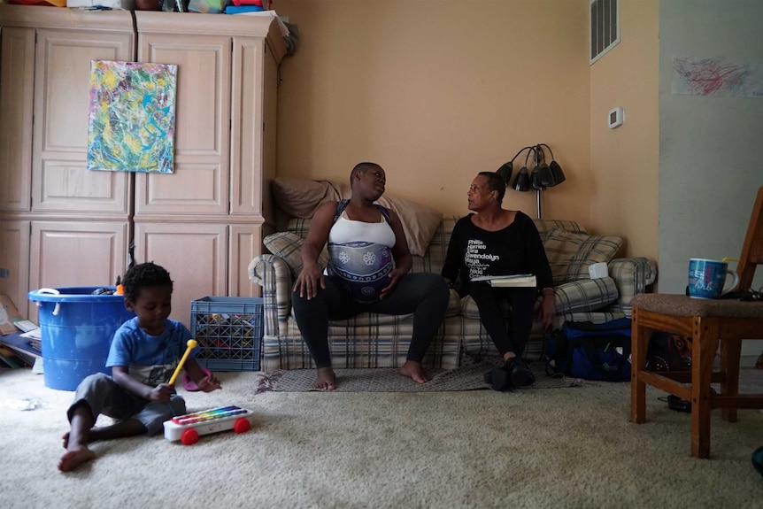 Binayake Joy and Claudia Booker sit on a couch and talk, while Binahkaye's toddler plays nearby.