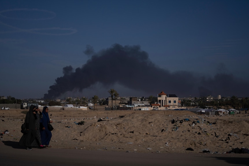 Smoke plumes in the distance as two people in hijabs walk in the foreground 