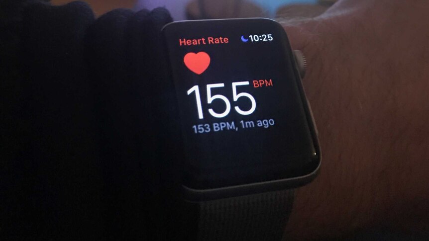 Picture of a heart monitor on a wrist, with a television in the background.