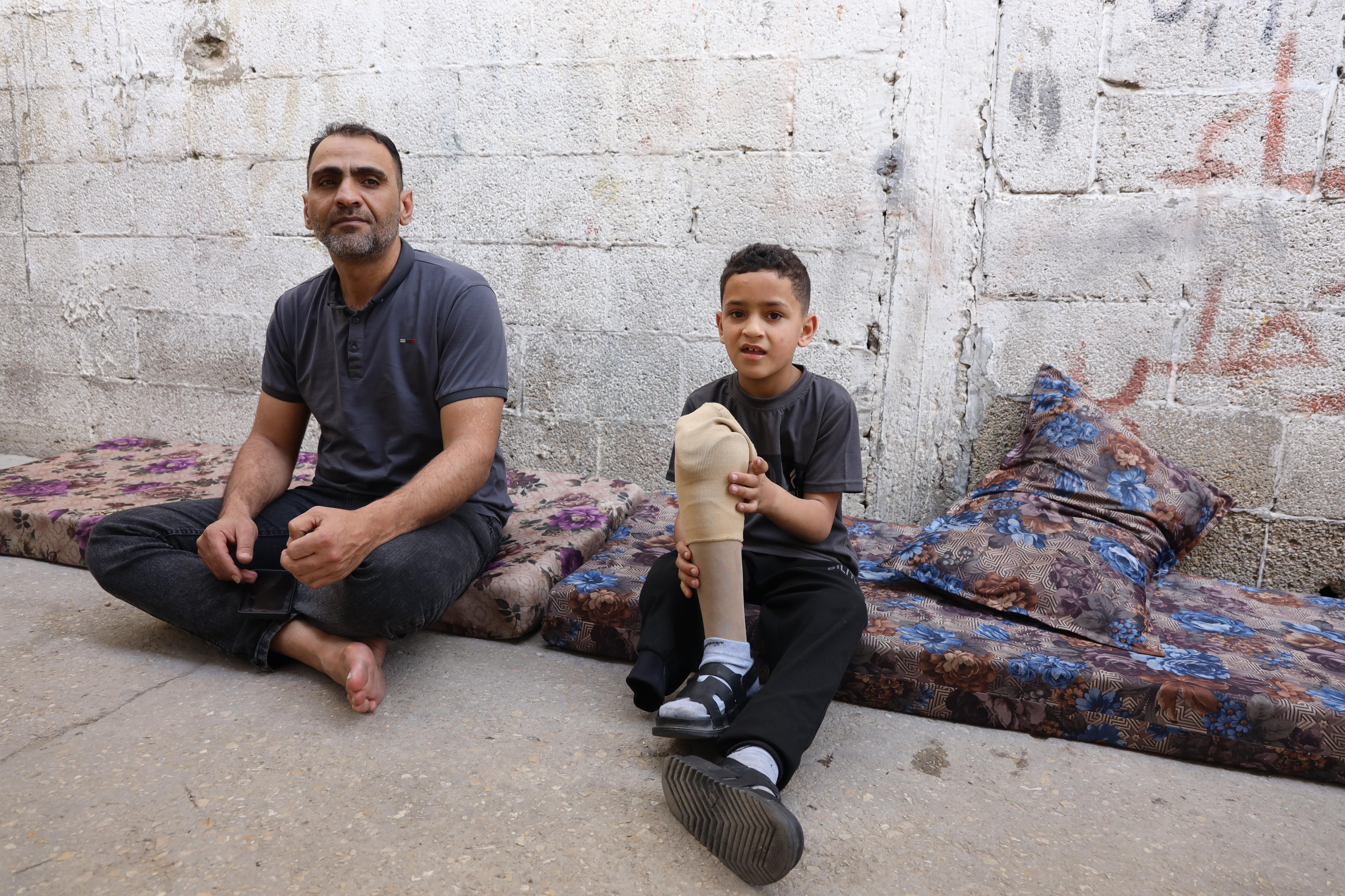 A man sits next to his son, who is holding a prosthetic leg, on a mattress on the ground.