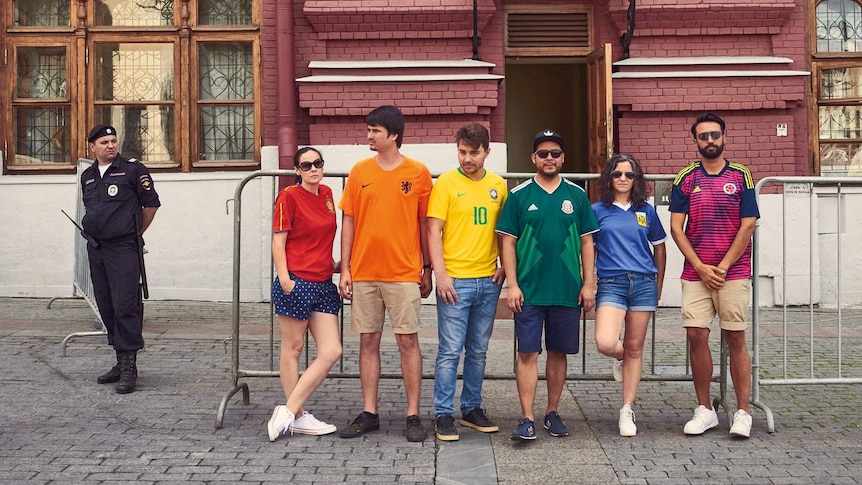 People wearing different coloured jerseys stand in a row in front of a red brick building.