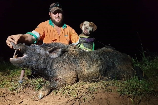 A pig hunter in hi-vis and his hunting dog sit behind a freshly-dispatched wild boar.