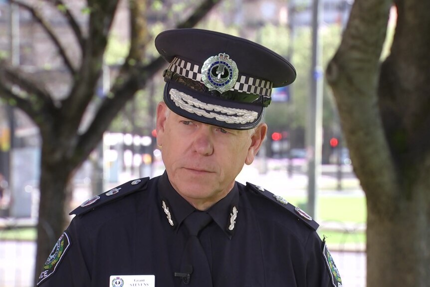 A man in police uniform looks off camera, with trees behind him