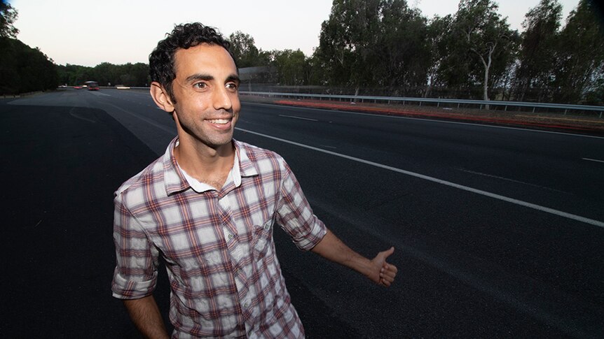 A man with dark hair, wearing a checked shirt, standing by the side of a highway.