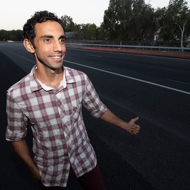 A man with dark hair, wearing a checked shirt, standing by the side of a highway.