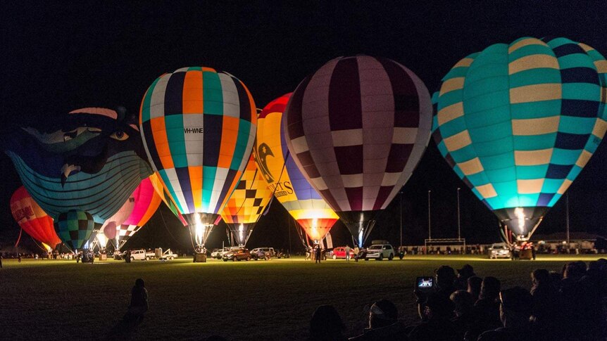 The jets of several hot air balloons light up the night sky as the balloons sit on the ground before take off.