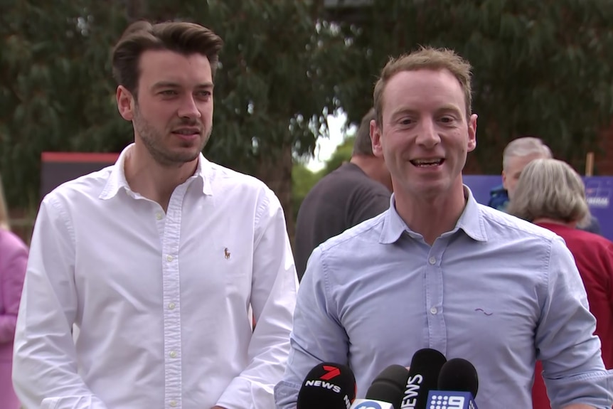 Two men wearing open-necked shirts speaking to microphones