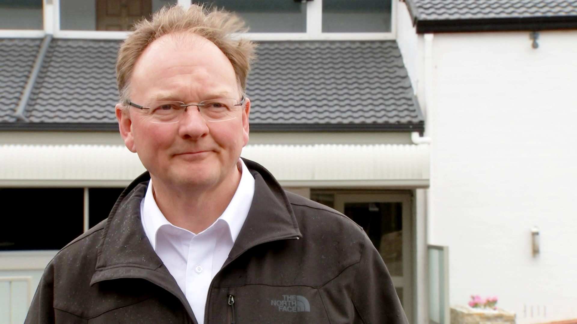 A balding man in glasses stands in front of a house