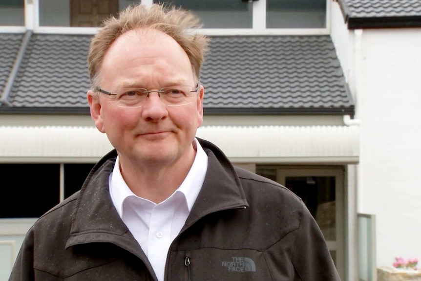 A bald man with glasses stands in front of a house