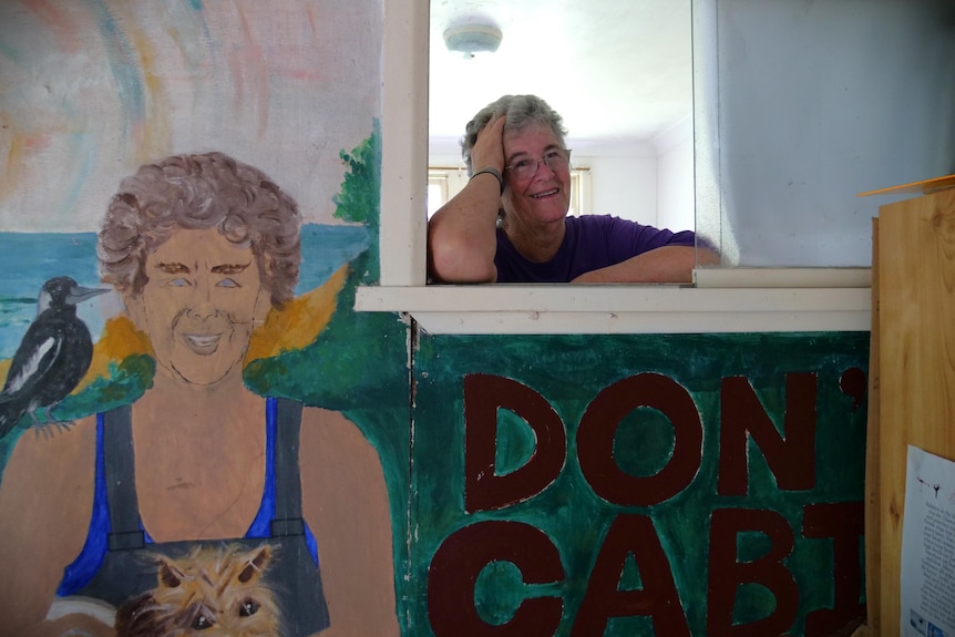 Woman with grey hair and purple shirt leaning into window, painting of Don Hearn on left-hand side.
