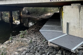 A perforated plastic walkway under a bridge, river nearby.